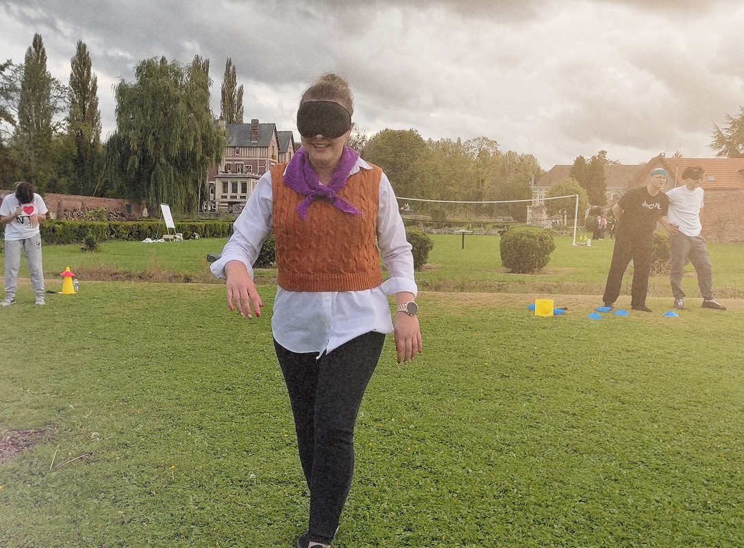 Une femme souriante marche les yeux bandés sur du gazon. D'autres participants à l'activité sont au fond, équipés de bandeaux.