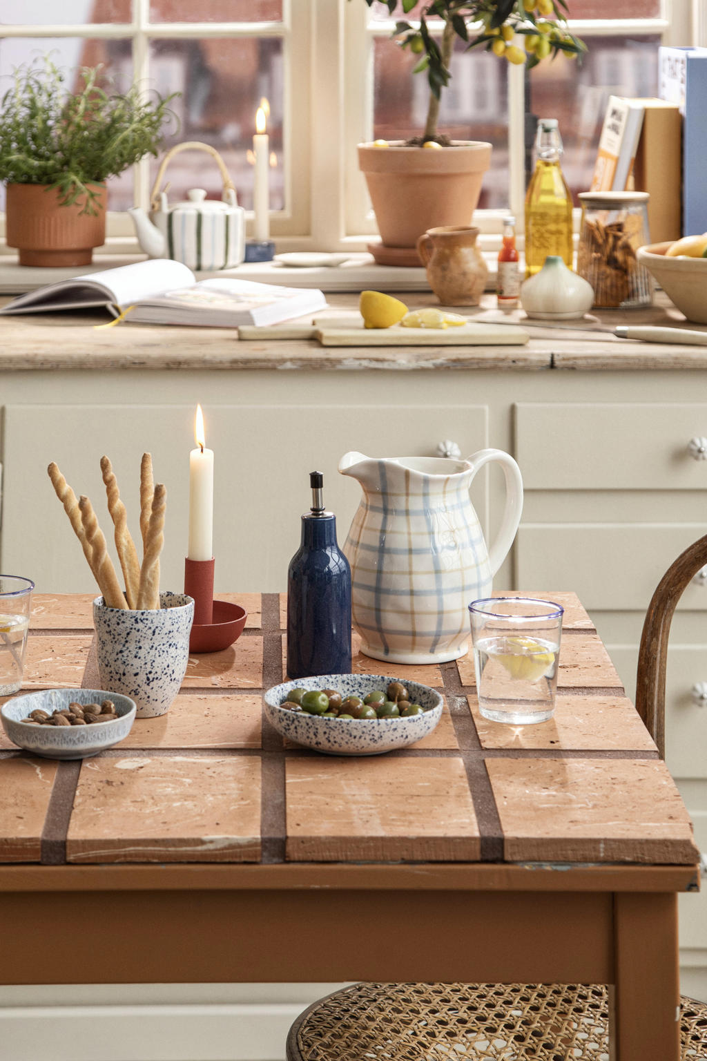 Table carrelée de style vintage avec bol et gobelet en céramique mouchetée bleu et blanc, pichet à carreaux, olives et grissinis, dans une cuisine lumineuse