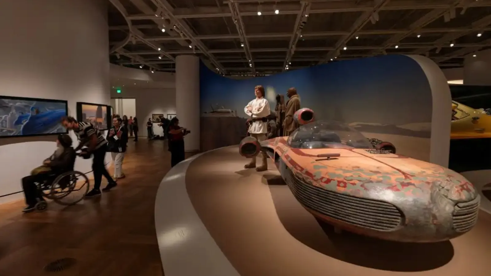 A museum exhibit features a detailed replica of a weathered, retro-futuristic vehicle resembling a hovercraft, surrounded by life-sized mannequins in a space-themed setting, under ambient lighting, with visitors exploring in the background.