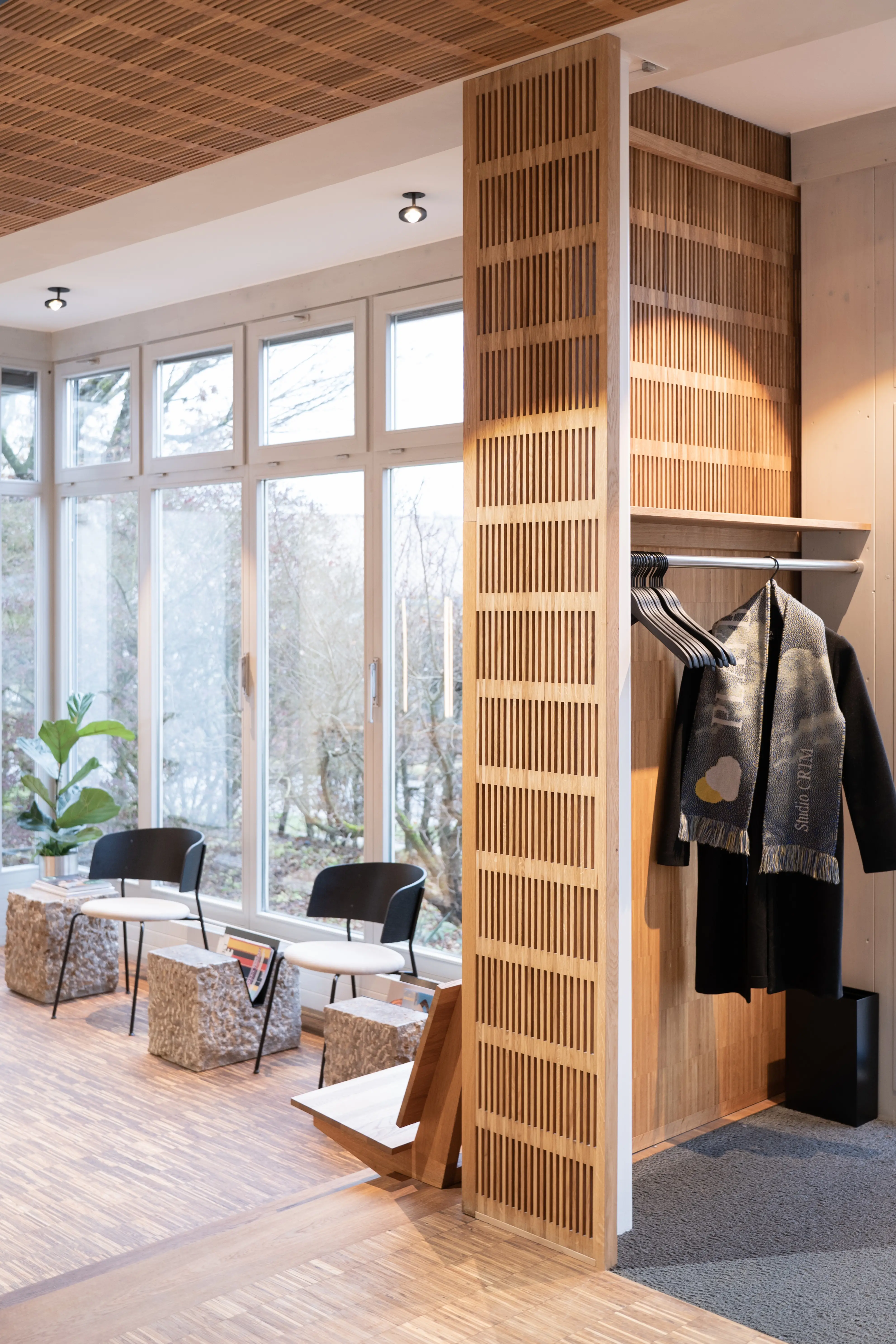 Waiting area at EMPL FRISEURE with black chairs and stone-look stools by the window front, a wooden slatted divider and an open wardrobe with coats.