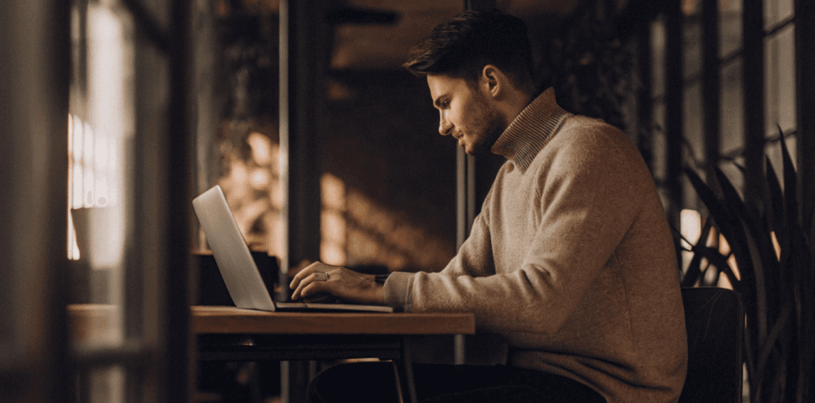 A man wearing a tan turtleneck sweater works on a laptop at a wooden table in a cafe