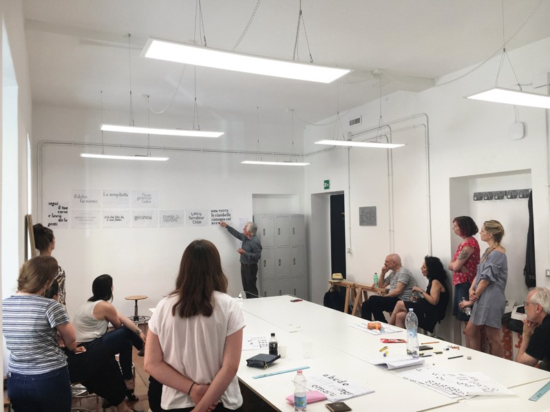 A photo of some students standing in a room while listening to the teacher talking about their letter drawings.