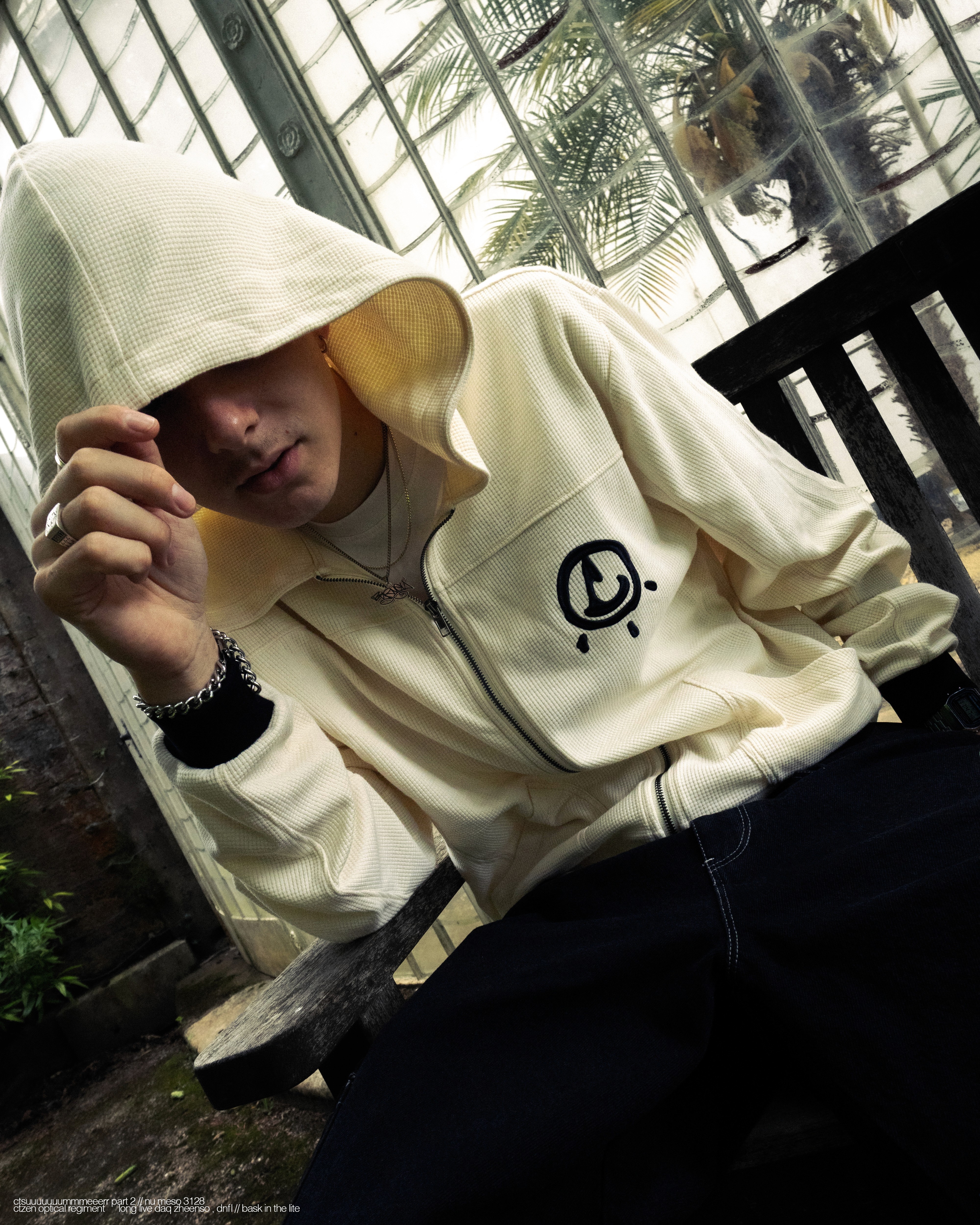 Smiling man in a white hoodie and black pants standing confidently on a bright green football field with empty stadium seats behind.