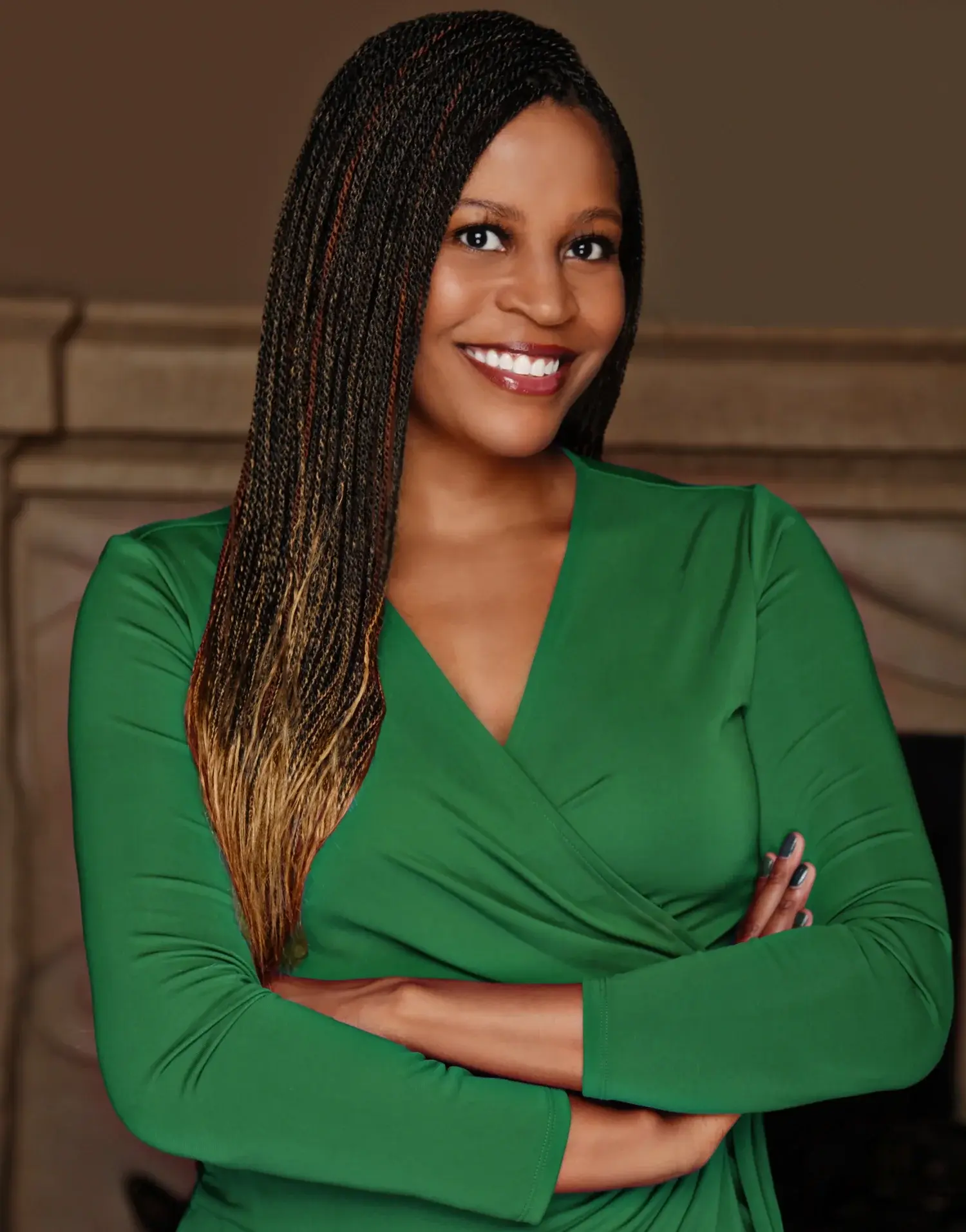 A woman with long, braided hair wears a green dress and smiles confidently with arms crossed against a neutral, indoor background.