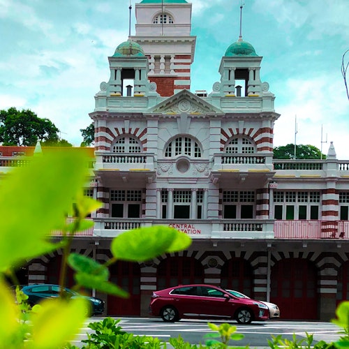 Historic white and red building with domed towers, cars parked in front, and foliage in the foreground.