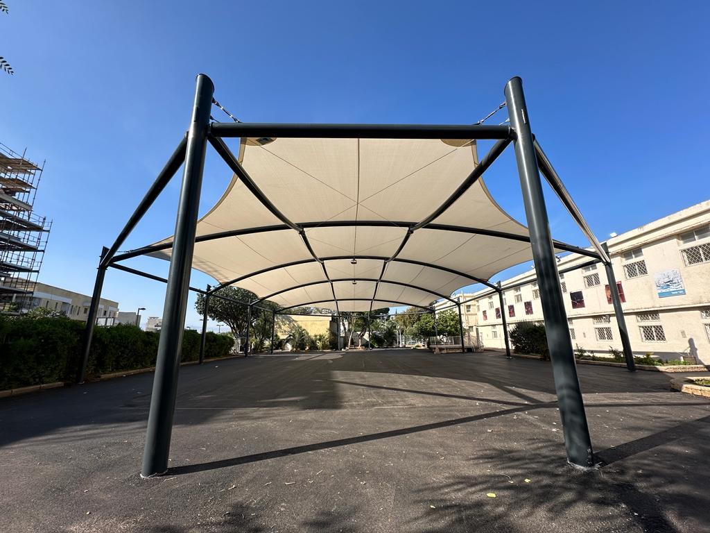 Large rectangular shade sail on black metal posts over an asphalt area in an urban setting.