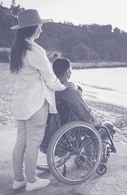 A woman in a hat standing next to a man in a wheelchair as they watch the sunset on a beach with a dog