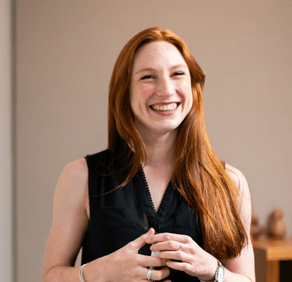 A woman with long red hair in a black sleeveless top smiles warmly. She stands in a softly lit room, conveying a cheerful and welcoming mood.