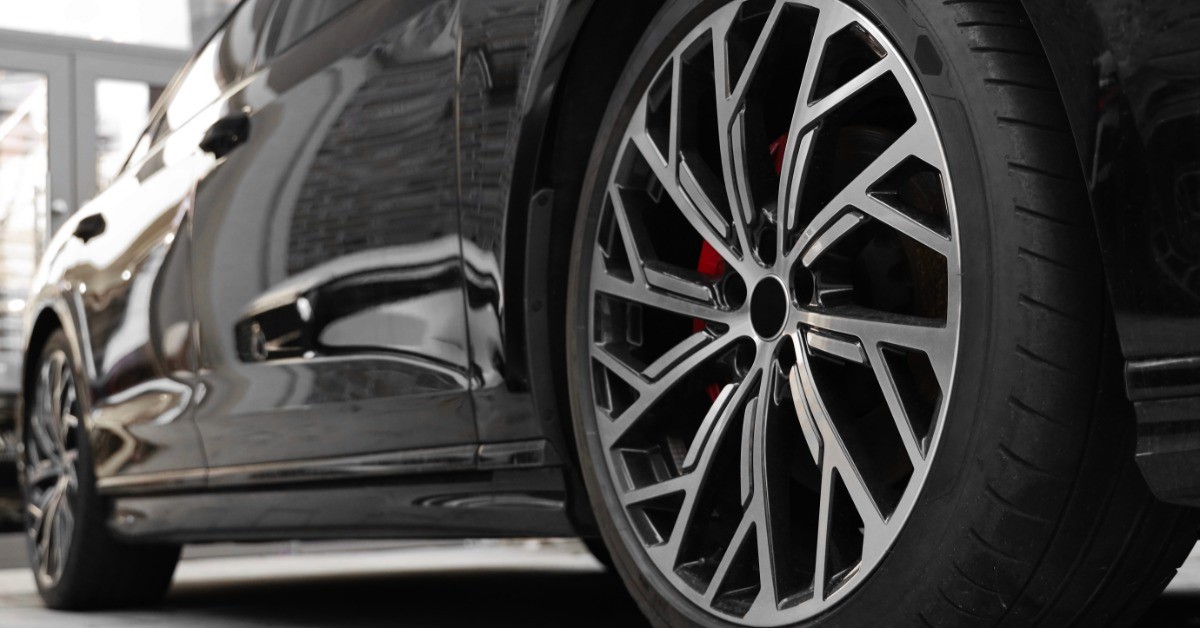 A black car with a large alloy wheel and low-profile tire parked on light paving inside a bright garage.