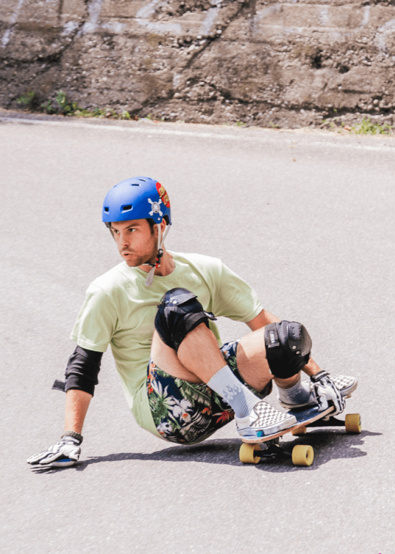 A person in a blue helmet and safety gear crouching low on a longboard while turning sharply on a smooth, downhill road. Graffiti is visible on a concrete wall in the background.