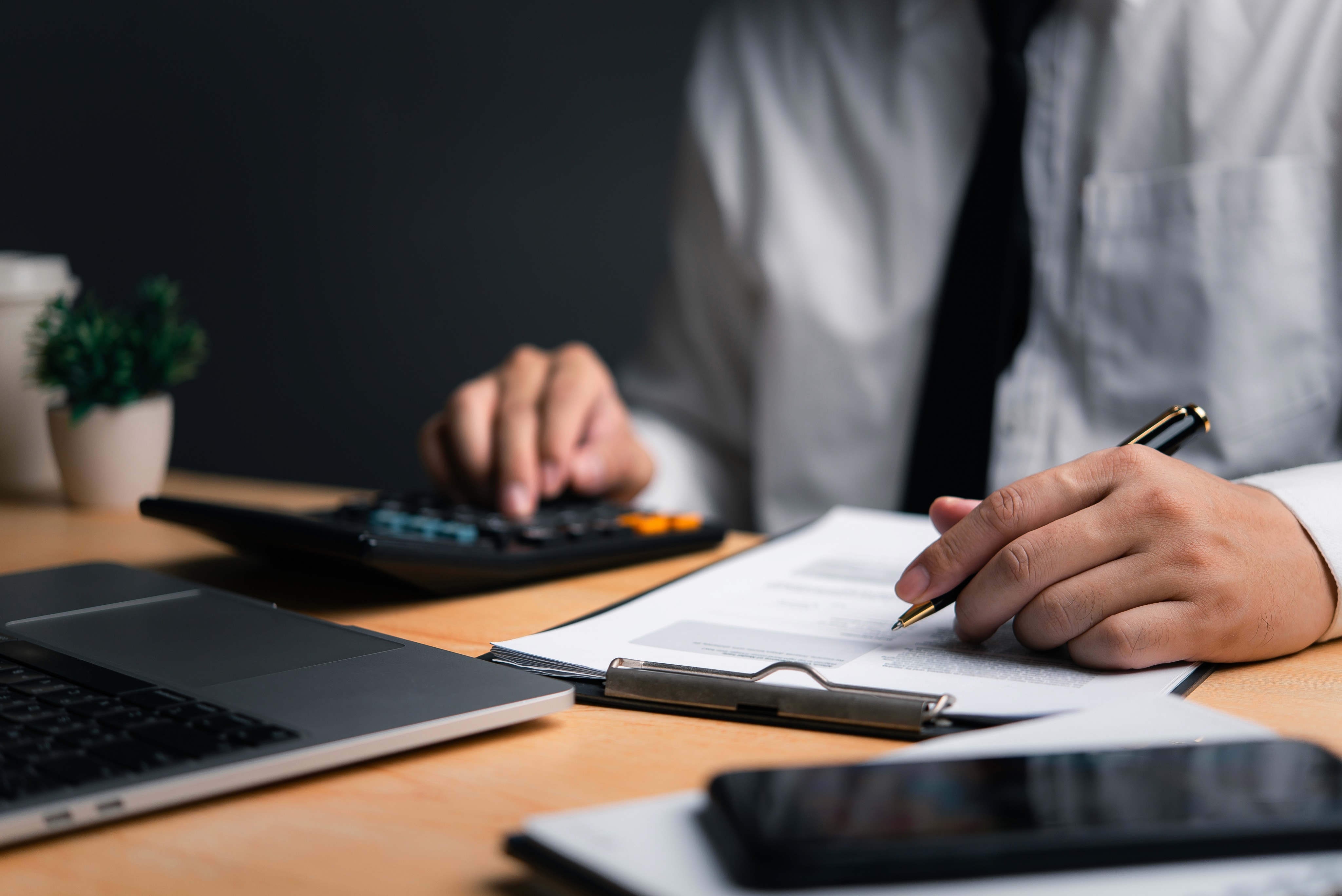 A person wearing a white dress shirt and black tie sits at a desk, using a calculator with one hand while holding a pen over a document with the other. 