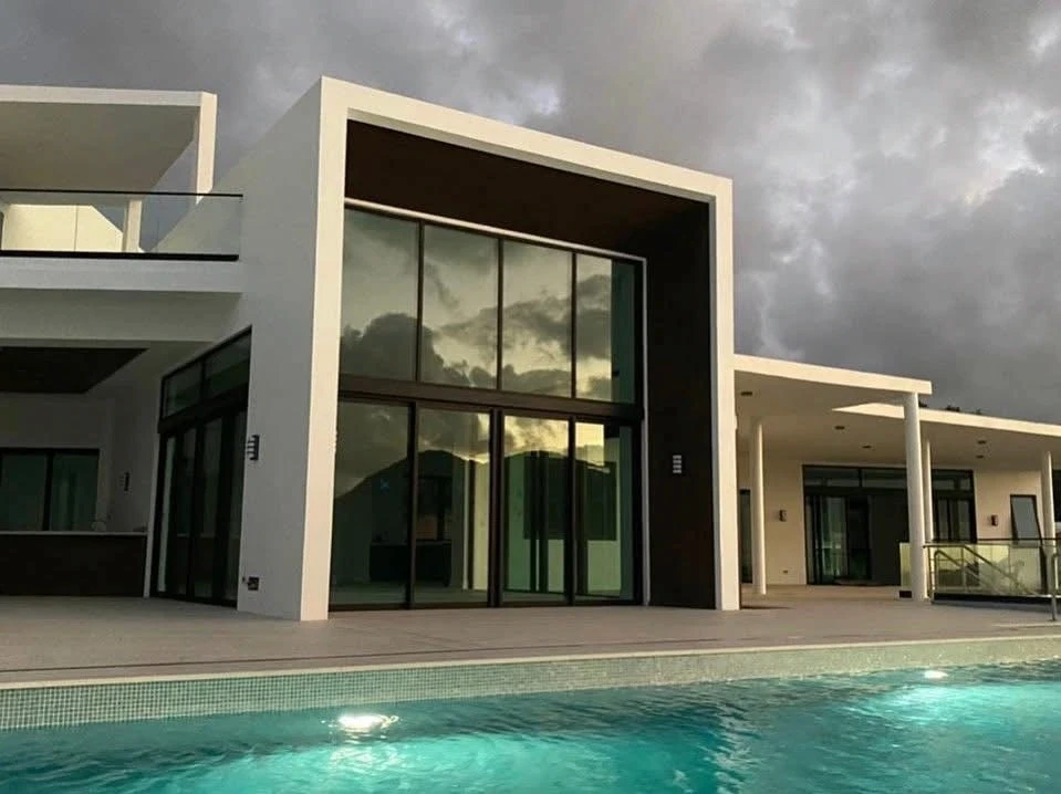 Double-height glass facade of The Hill Residence in St. Lucia by Atelier International, viewed across the pool at dusk under a stormy sky.