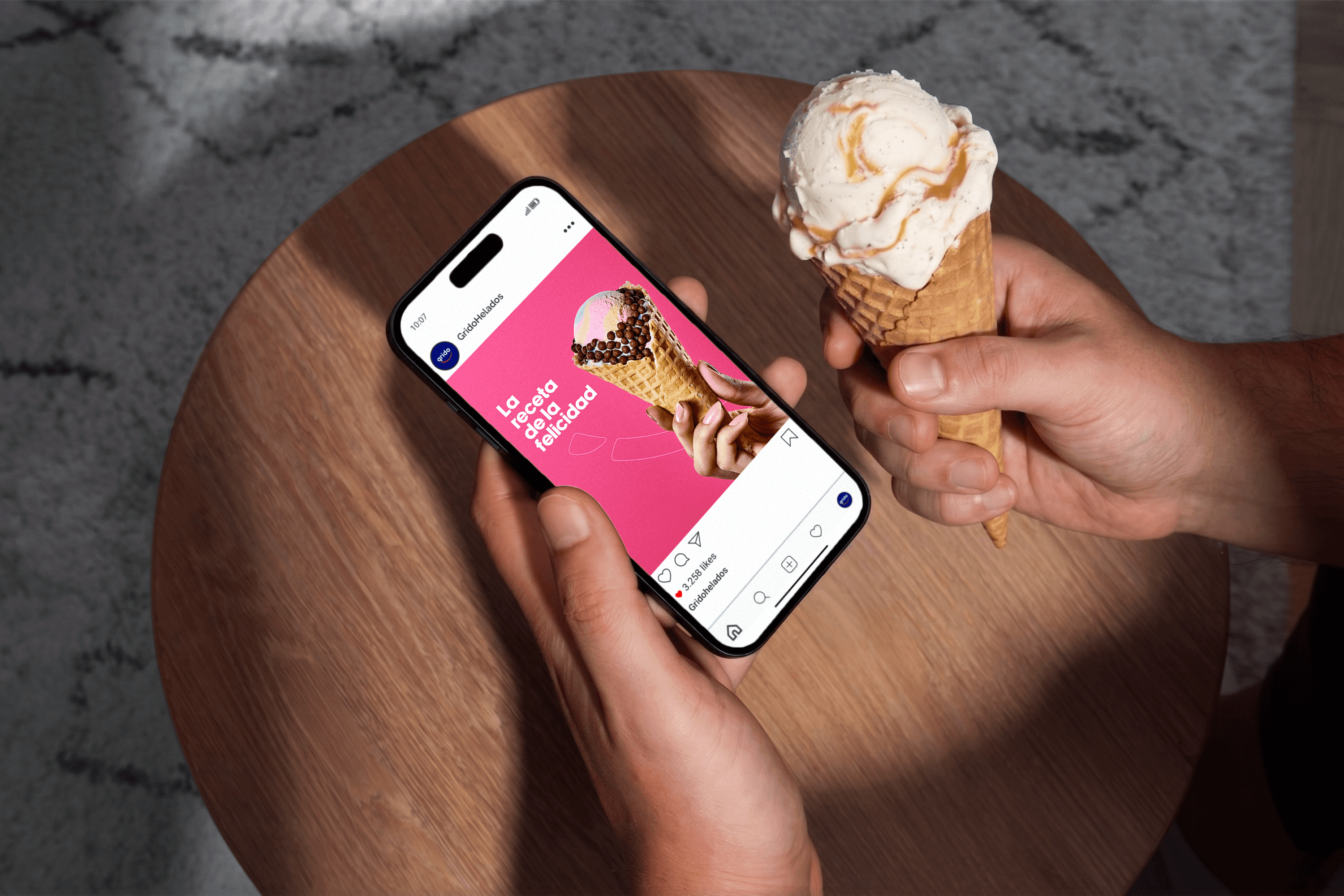 Top-down view of a person holding a waffle ice cream cone in one hand and a smartphone displaying a pink Grido Helados Instagram post in the other over a round wooden table.
