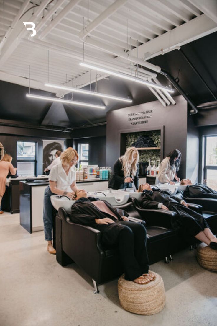 Stylists wash hair at modern stations in an industrial salon with dark walls and exposed ceiling pipes.