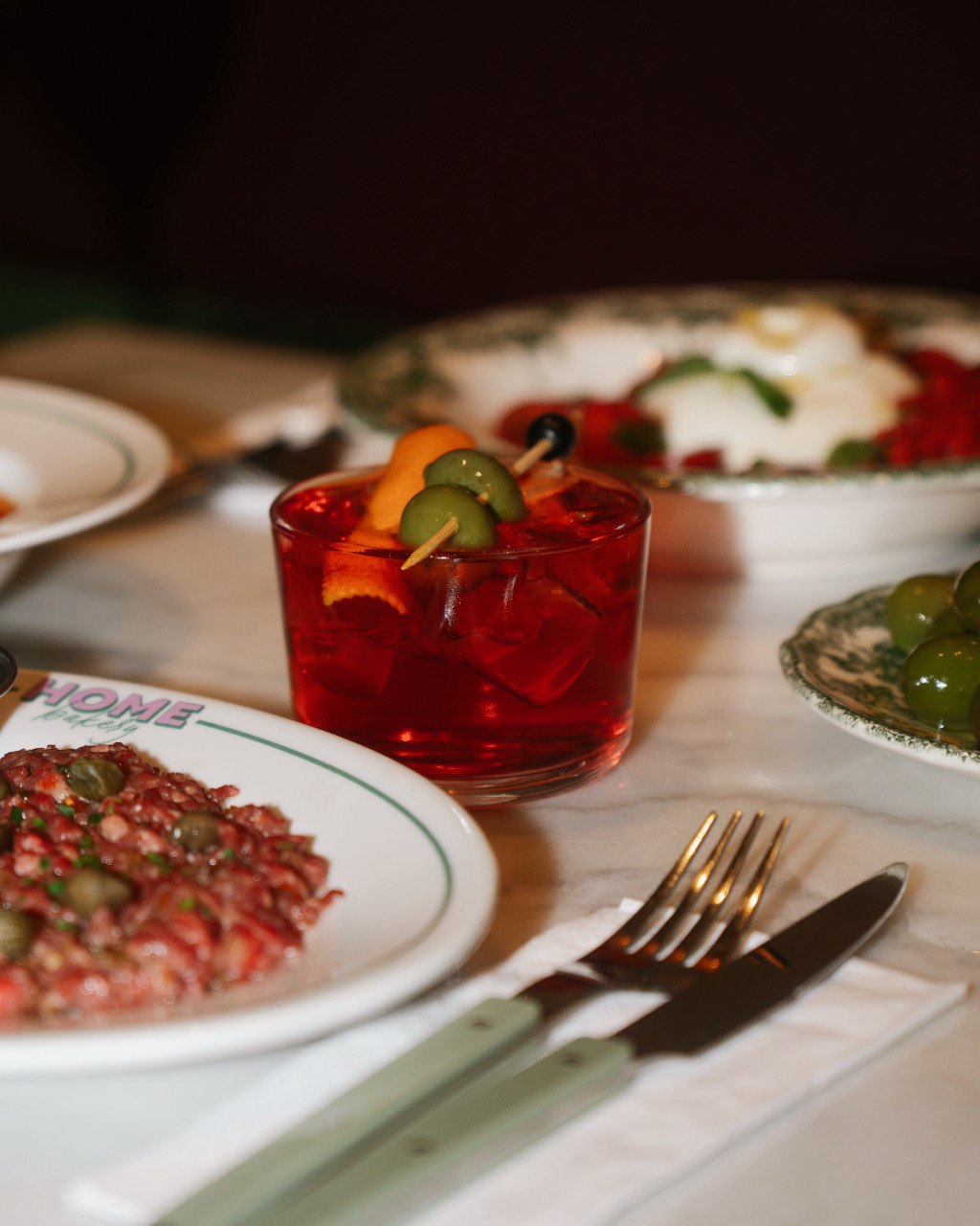 A luxurious dining table set with a vibrant red cocktail garnished with orange peel and olives, surrounded by elegantly plated dishes including a meat dish topped with capers and a fresh green olive salad.