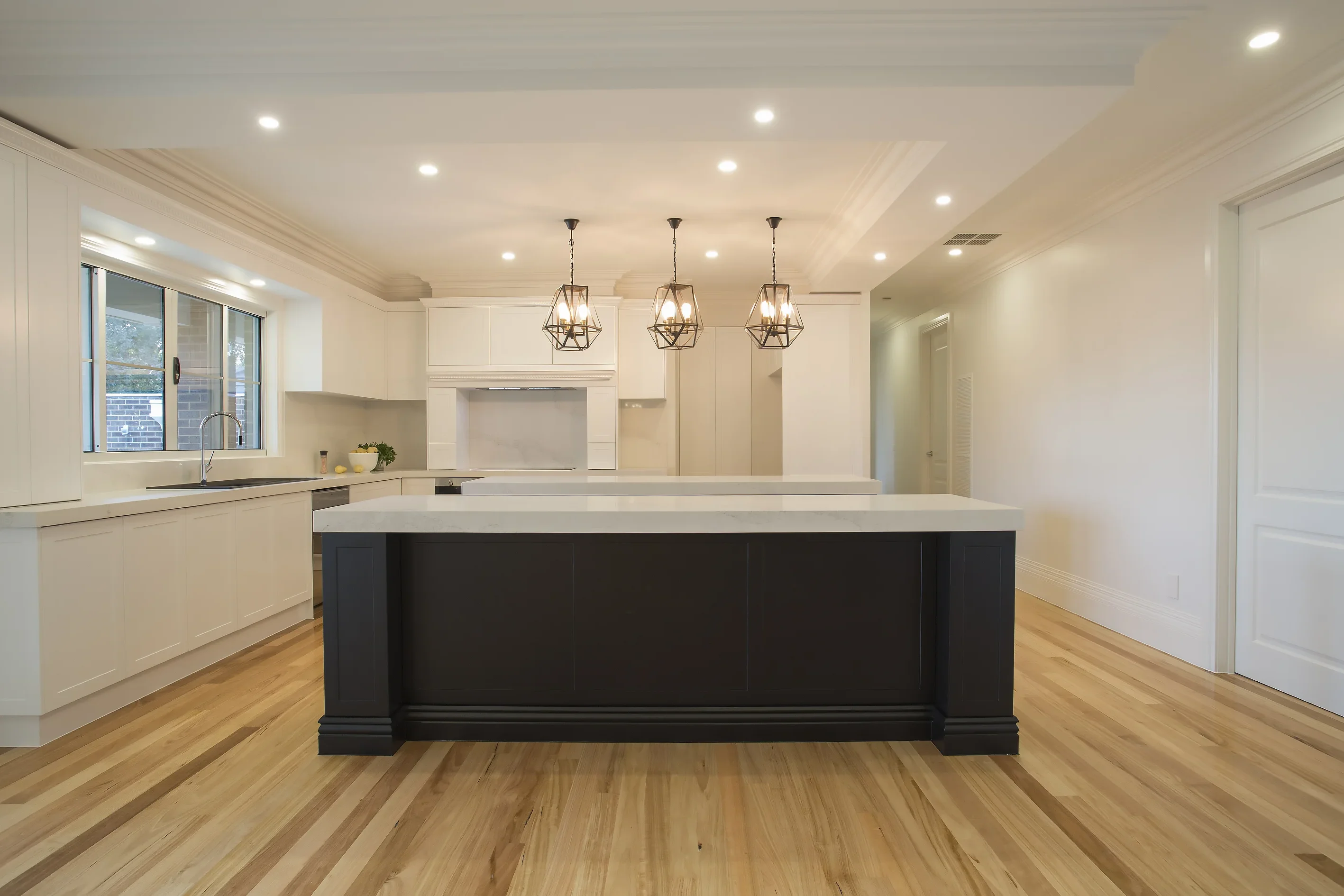 Kitchen viewed head-on with a black island bench, white stone benchtop, three caged pendant lights and white cabinetry