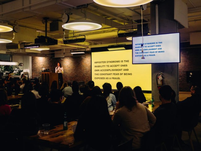 Referentin spricht vor einem Publikum in einem abgedunkelten Veranstaltungsraum, im Hintergrund eine beleuchtete Präsentationsleinwand