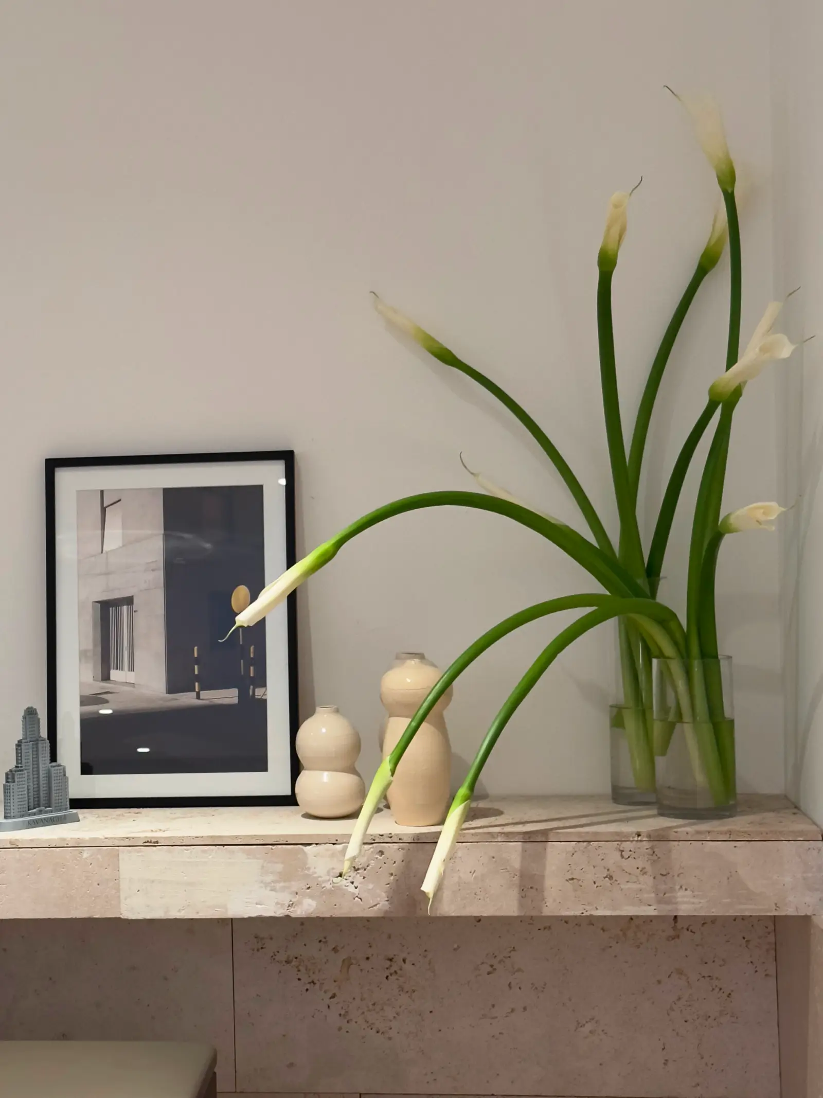 Rincón decorativo con calas blancas en florero de vidrio, cuadros de arquitectura y piezas de cerámica neutras.