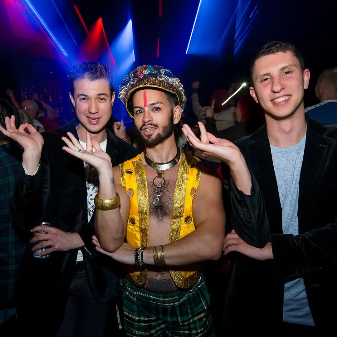 Three people pose at a nightclub with colourful lights. The person in the centre wears vibrant, ornate clothing and jewellery, while the two on either side wear dark jackets. All three hold their hands up in a similar playful gesture.