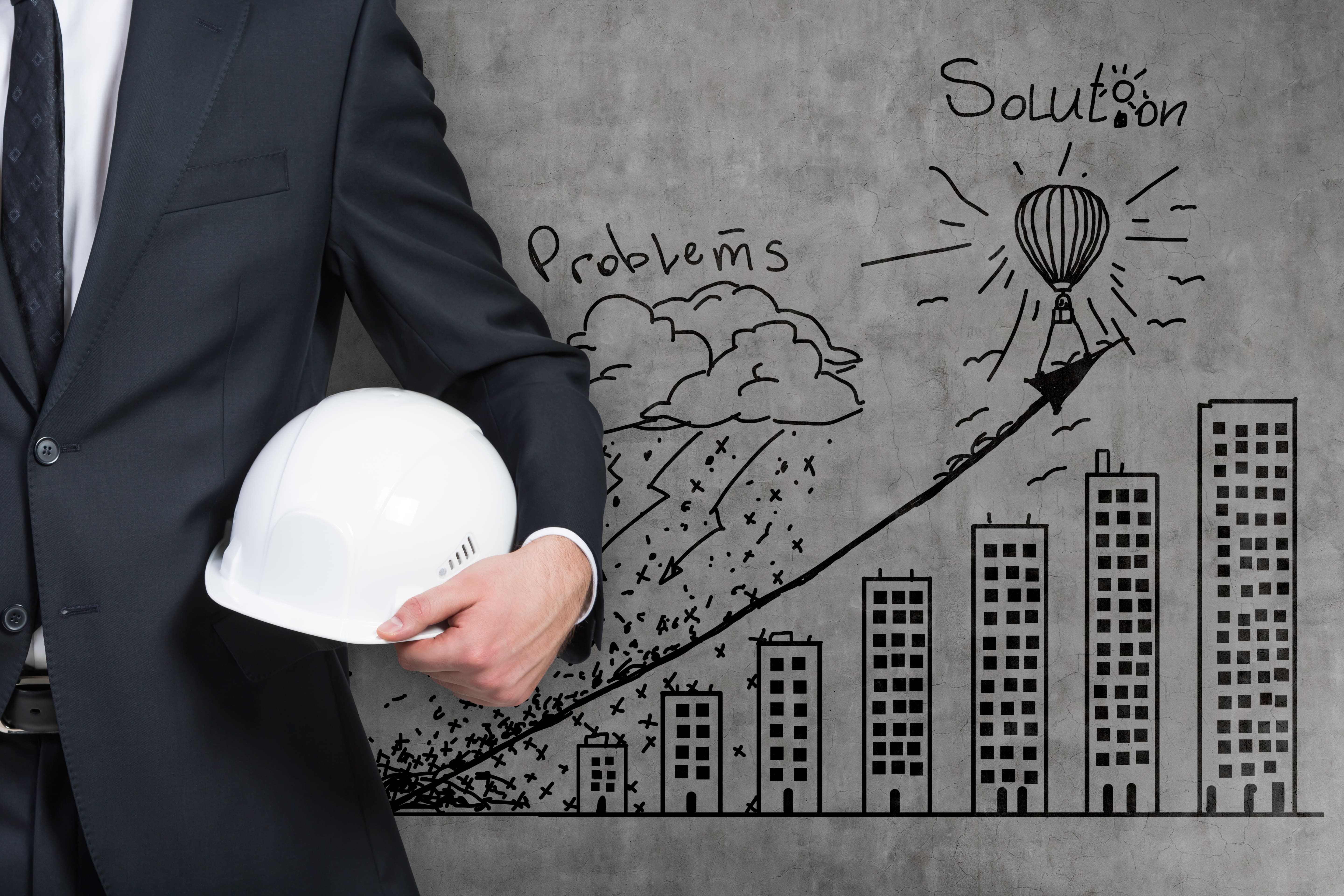 A man in a suit holding a white hard hat, standing in front of a wall with a business-themed sketch showing “problems” leading to “solutions.