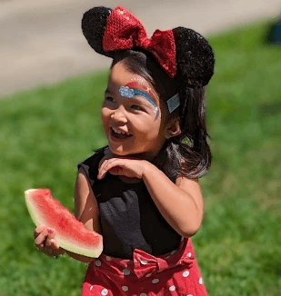 Smiling young girl with a Minnie mouse outfit, glittery bow and ear headband, and rainbow face paint enjoying a watermelon