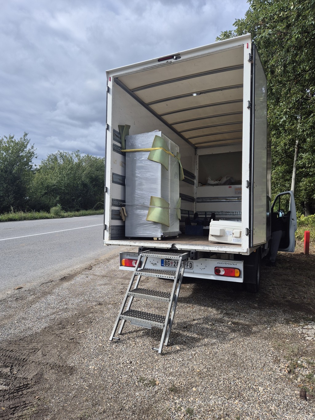 Kamion za selidbe parkiran pored puta, sa otvorenim tovarnim prostorom u kojem je uredno upakovana bela tehnika obmotana zaštitnom folijom i kaiševima, spremna za transport.