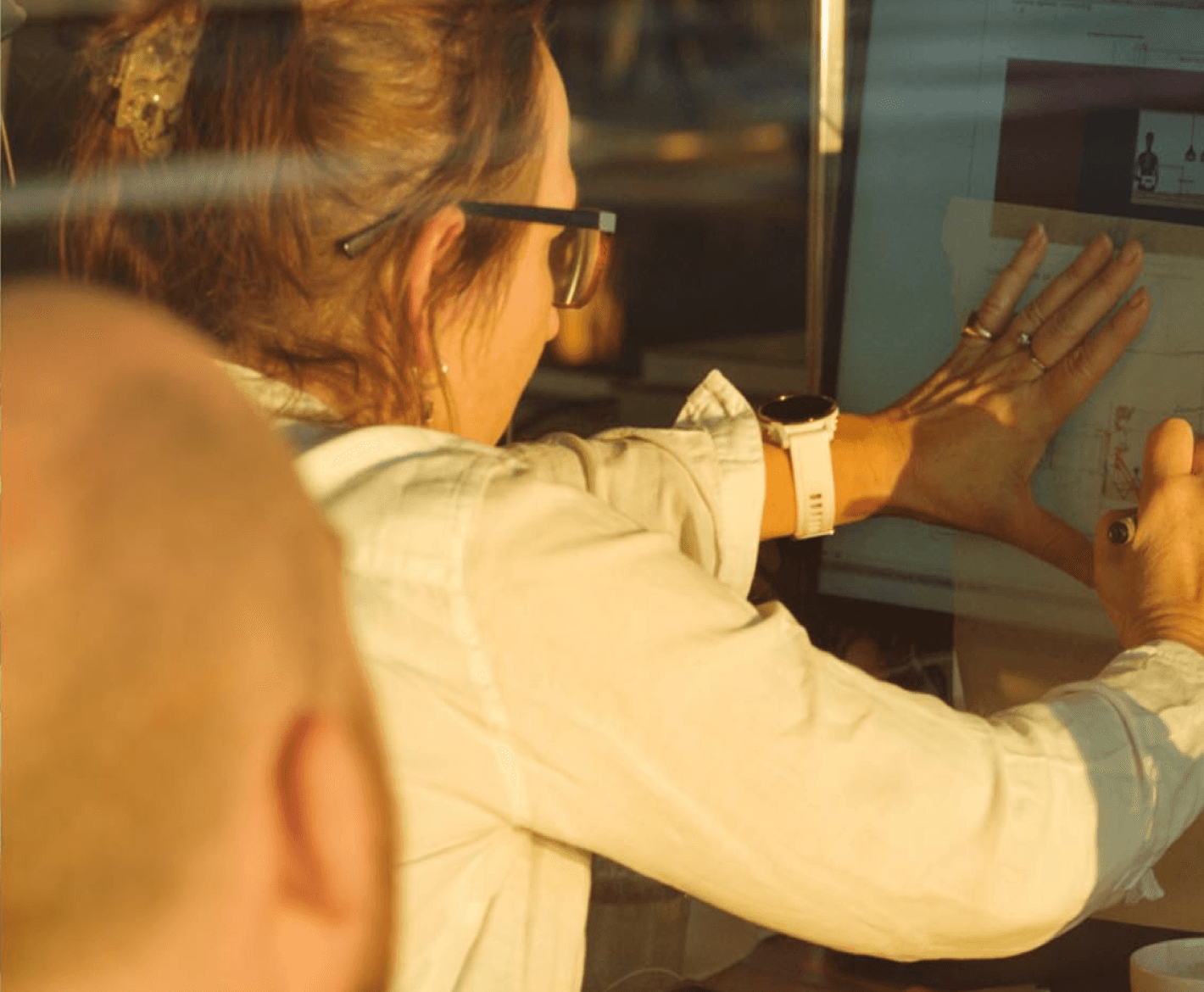 a close-up of a woman interacting with a screen, possibly presenting or explaining something. She wears glasses and has her hand raised, emphasizing her point.