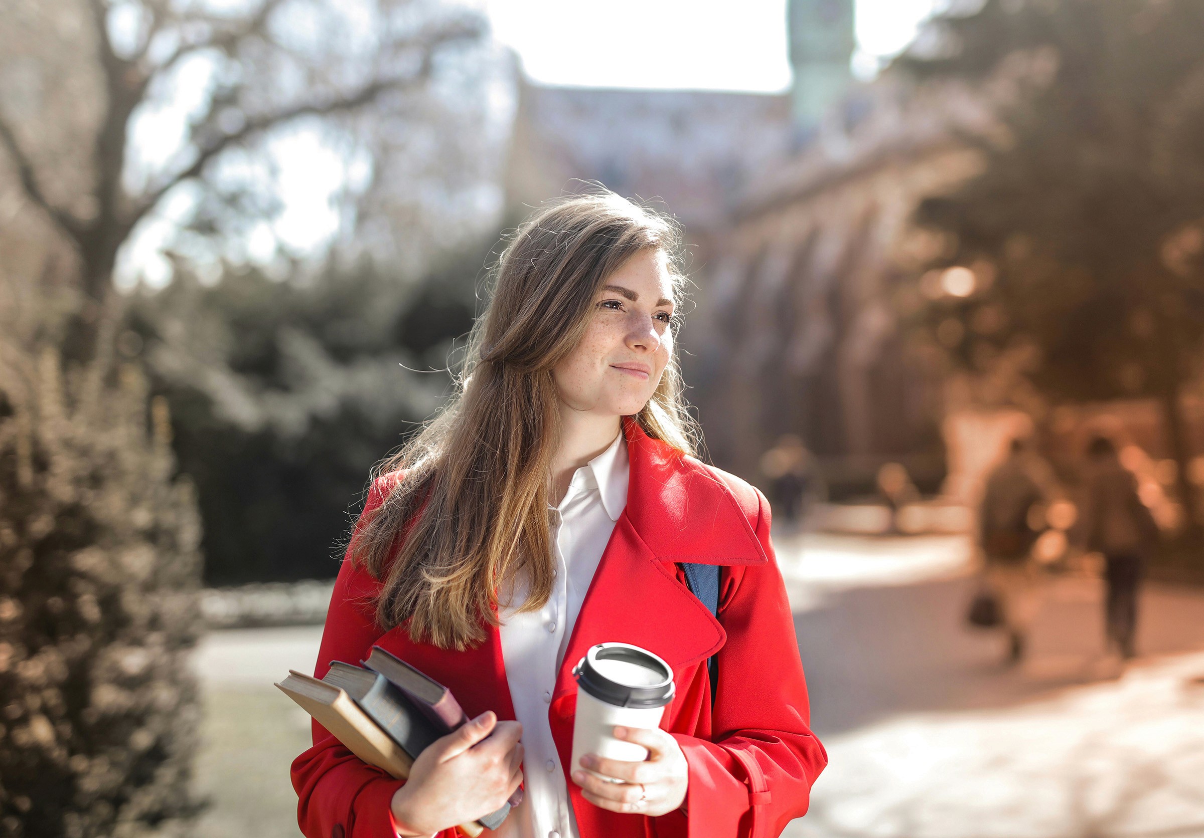 A schoolgirl stands outdoors holding books in one hand and a coffee cup in the other.