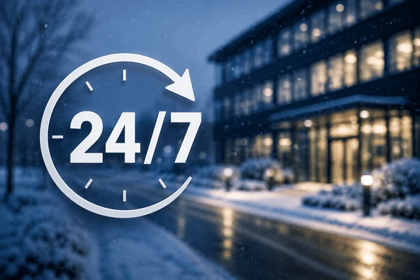 Ein modernes Bürogebäude bei Dämmerung im Winter mit einem großen, halbtransparenten „24/7“-Grafiksymbol im Vordergrund, das für einen Rund-um-die-Uhr-Service steht.