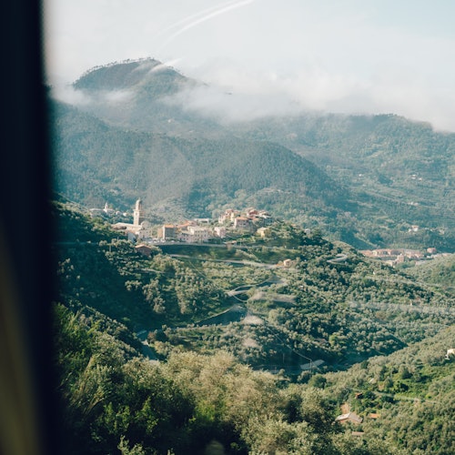 Aerial view of a small village with a prominent tower nestled in lush green hills and valleys under a partly cloudy sky.