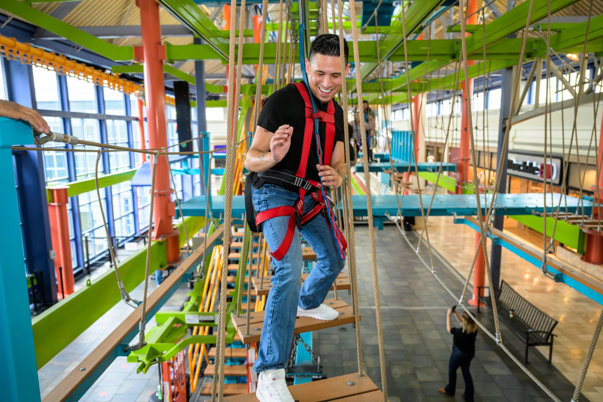 Guest in a safety harness crossing a suspended plank on the LiggettVille ropes course above the mall floor