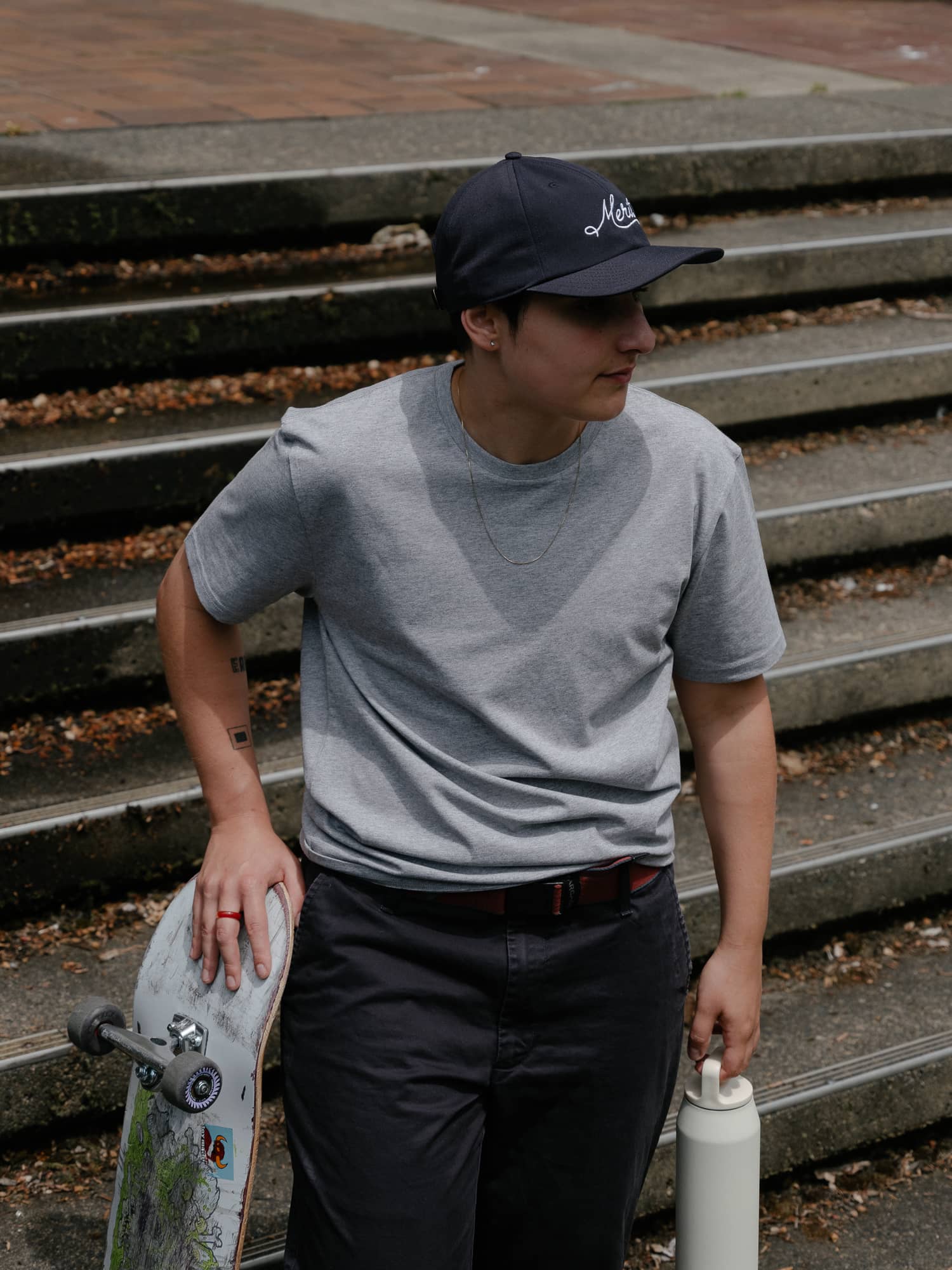 Young person in a gray t-shirt and navy cap holds a skateboard and water bottle. Standing near concrete steps, they appear relaxed and thoughtful.