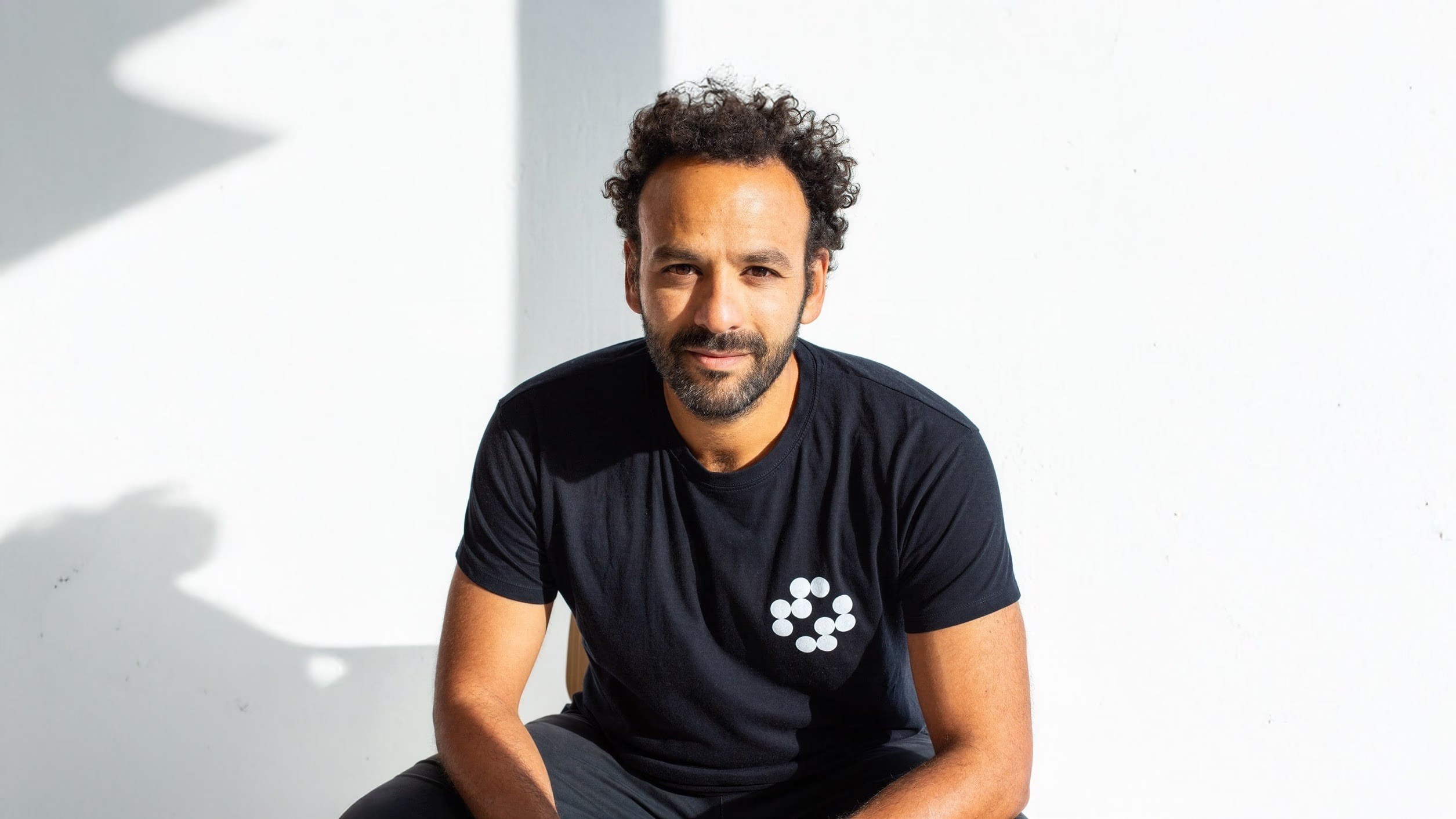 Een man met donker krullend haar en baard draagt een zwart T-shirt met wit patroon. Hij zit voor een witte muur met schadu...