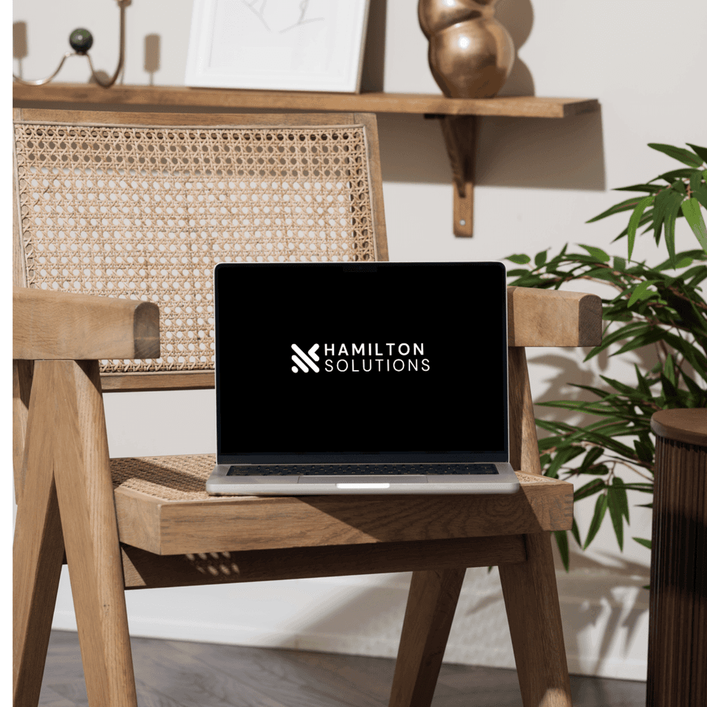 An image of a macbook resting on the wooden chair showing the Hamilton Solutions logo.