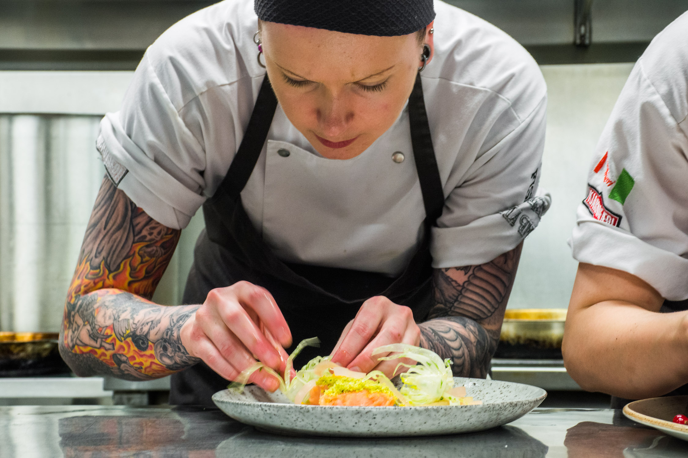 A chef with tattoos and a black headband plates a gourmet dish featuring seared fish and vegetables in a professional
