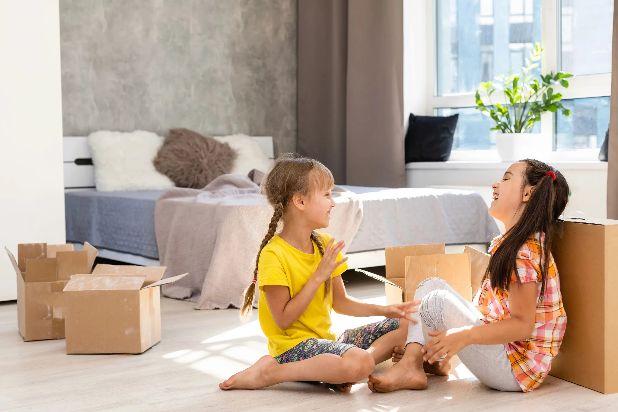 Two girls laughing, with open moving boxes behind them in the bedroom.