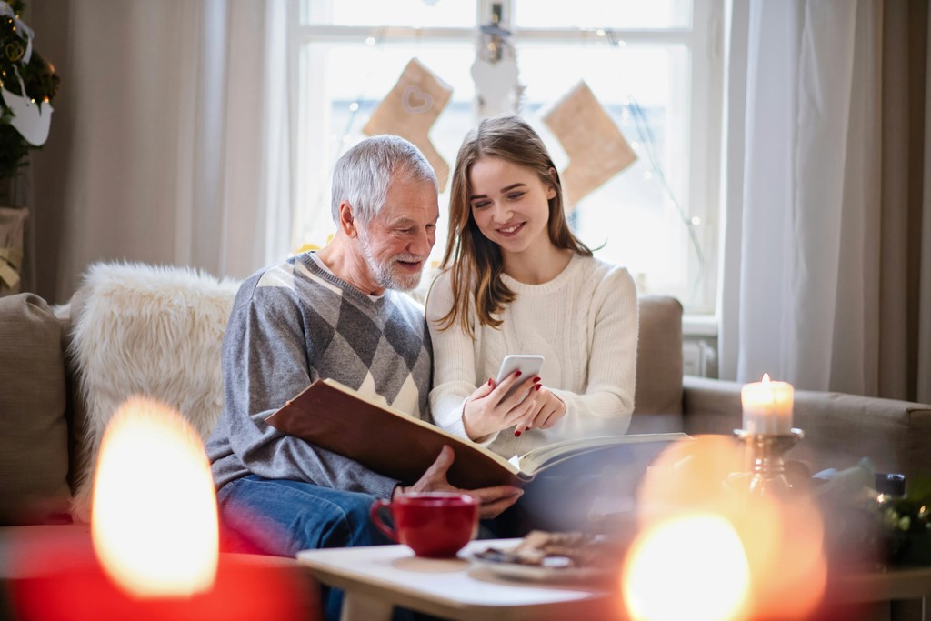 A grandfather and his granddaughter share a cozy moment in a warmly lit living room, flipping through a photo album and looking at a smartphone together. The festive atmosphere is enhanced by lit candles and holiday decorations, emphasizing family bonding, storytelling, and the joy of sharing memories across generations.