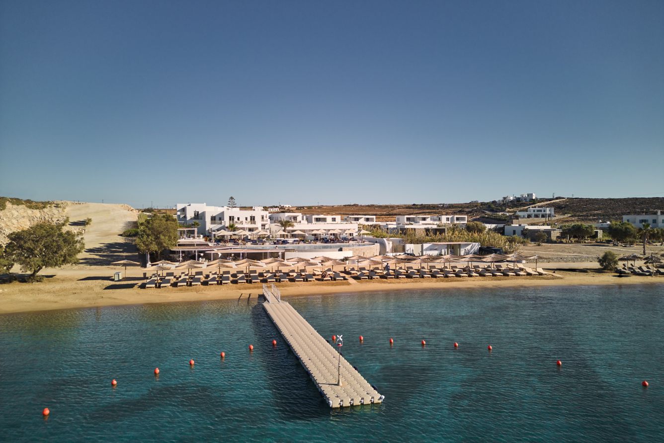 Aerial view of a beach resort with white buildings, many parasols, and a long pier extending into the clear, blue water.
