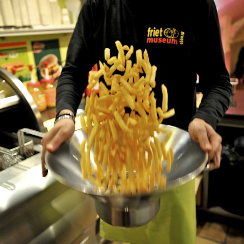 Személy, aki egy tállal dobálja a sült krumplit a Friet Múzeumban. A háttérben konyhai felszerelések és színes poszterek láthatók.