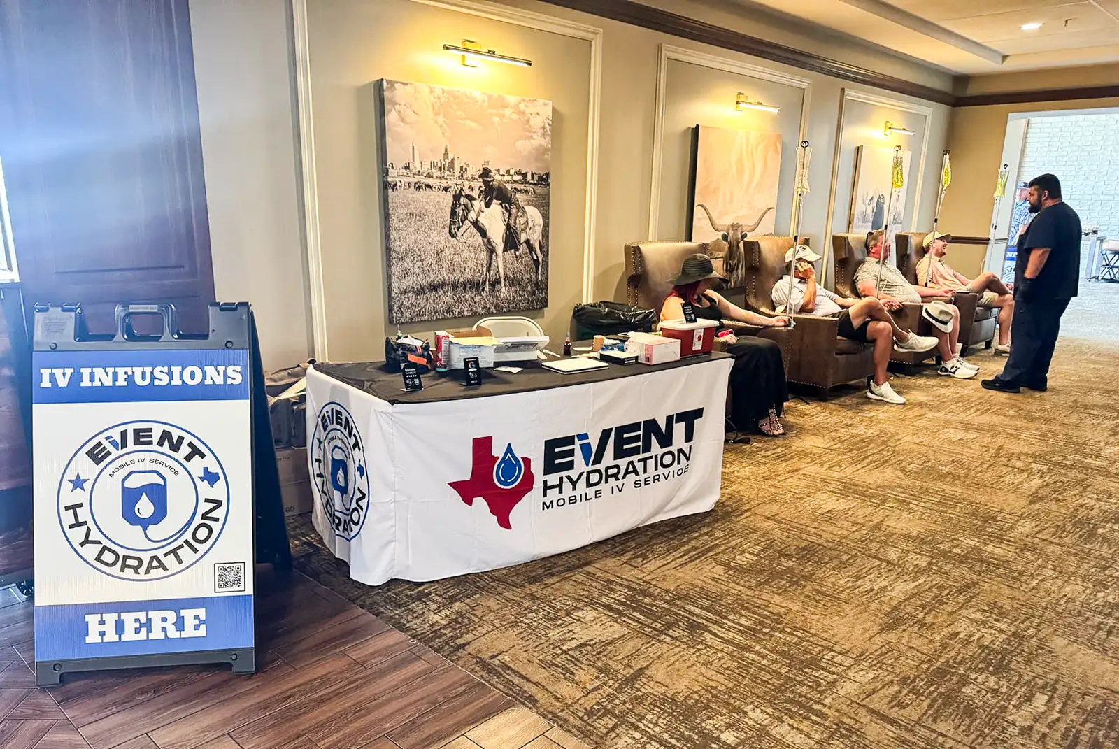 A group of guests receiving IV drips while seated in leather lounge chairs at an Event Hydration mobile IV setup in a hotel event space, with an "IV Infusions Here" sign and branded table