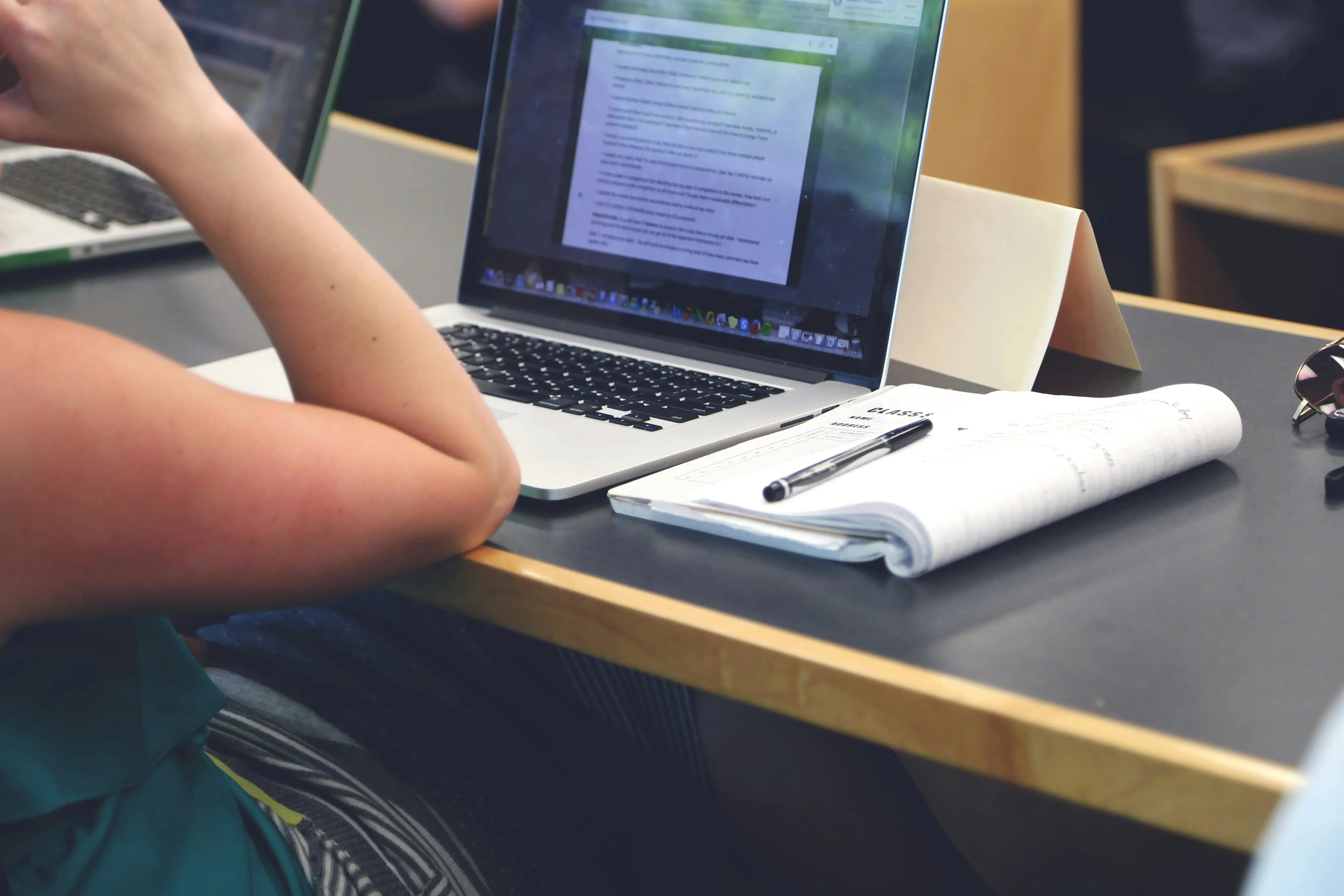 photo of open laptop with a notebook and pen next to it on a dek. there's an arm propped on the desk
