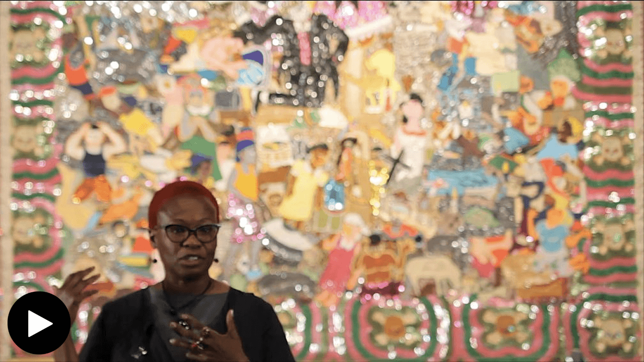 Gina Athena Ulysse stands in front of "Haiti Madi 12 Janvye 2010." Ulysse's hands are slightly raised in front of her as she speaks. The flag behind her is slightly out of focus, shimmering in the light.