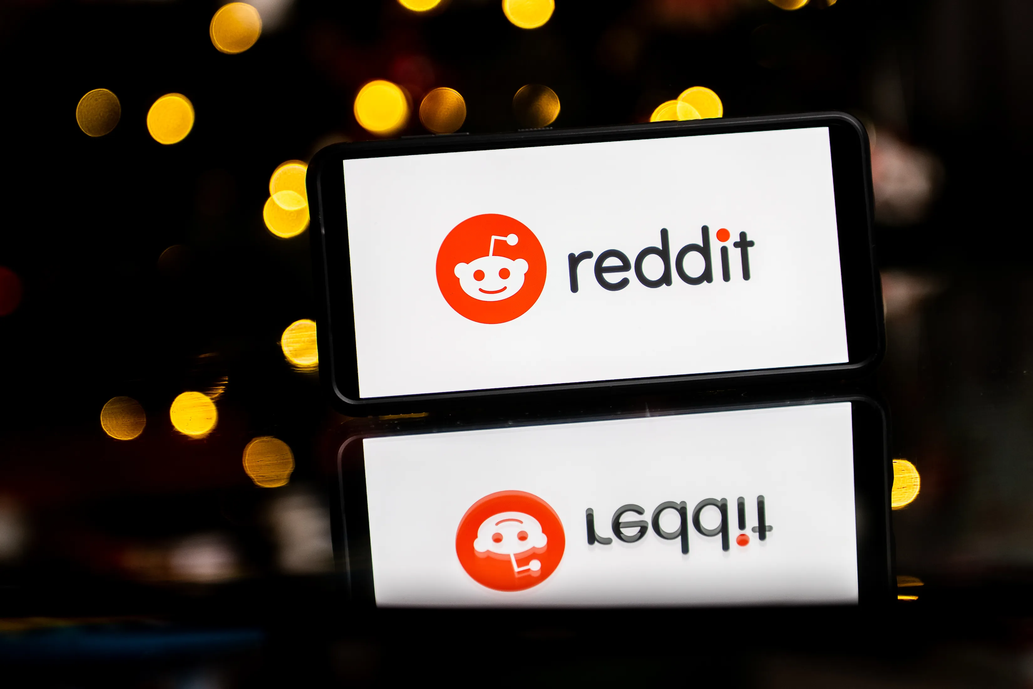 Reddit logo displayed on a smartphone screen with a reflection, set against a dark background with blurred yellow bokeh lights.