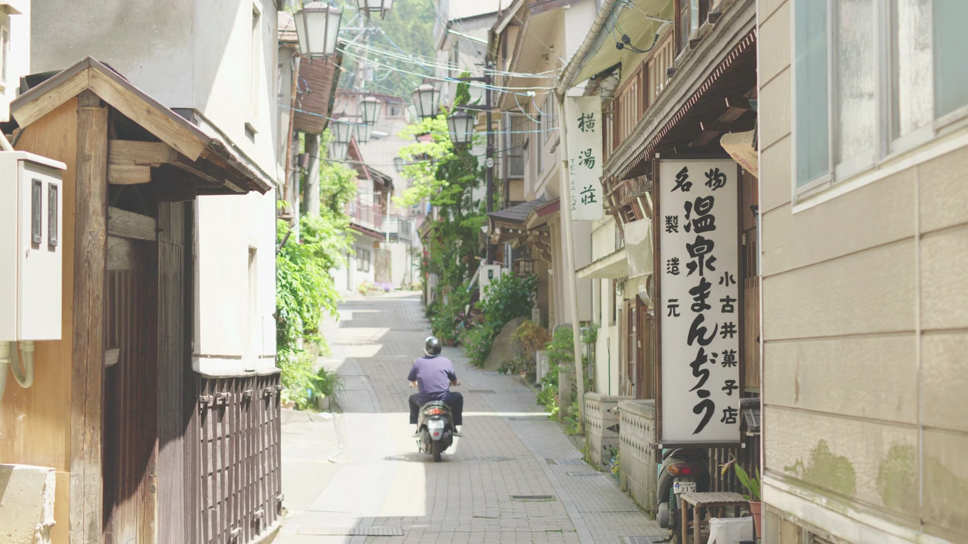 A local rides a scooter down a Shibu Onsen lane past an onsen-manjū (steamed bun) shop and hanging lanterns.