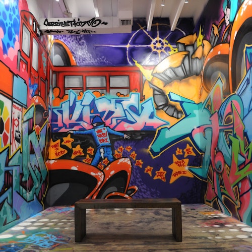房间的墙上满是彩色的涂鸦艺术，展现了抽象的设计和形状，中央放置着一条木凳，地板显得有些磨损。