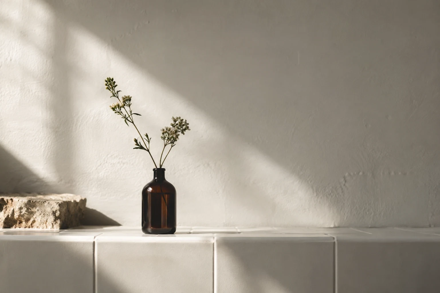 Single small dark glass bottle on a white tiled wall ledge, large negative space and delicate diagonal sunlight