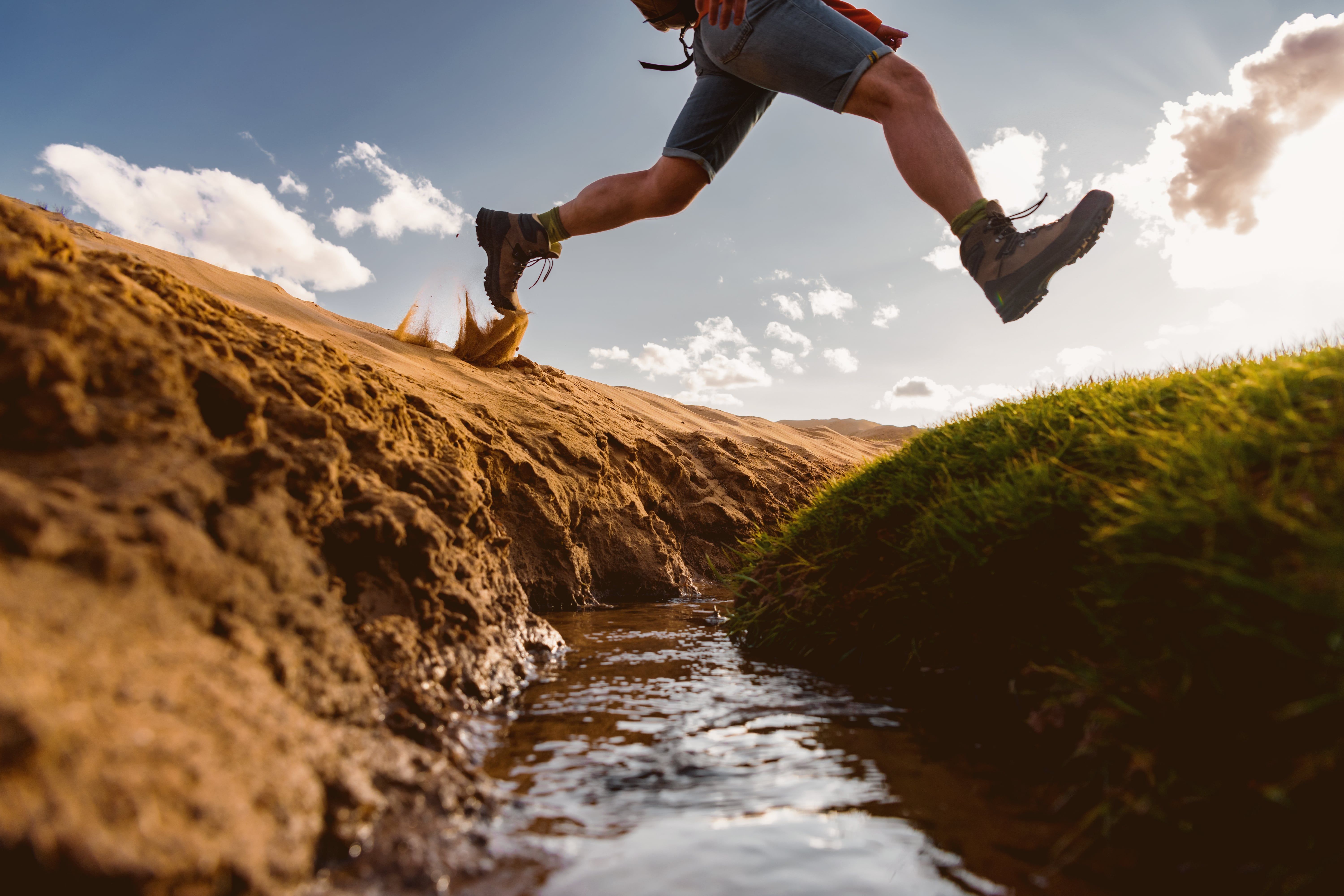 A person wearing hiking boots and shorts mid-jump over a small stream between a sandy terrain and a grassy mound