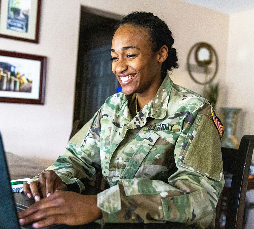 A woman in a U.S. Army uniform smiles while using a laptop.