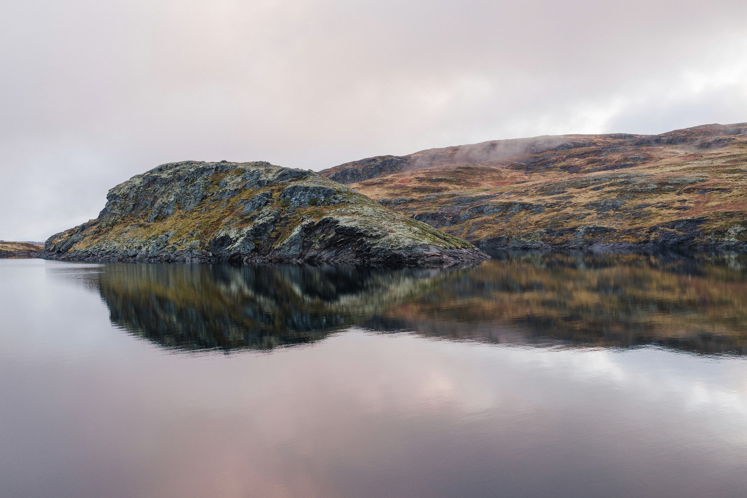 Minimal rocky shoreline and low mountain reflected in still water beneath a pale sky.