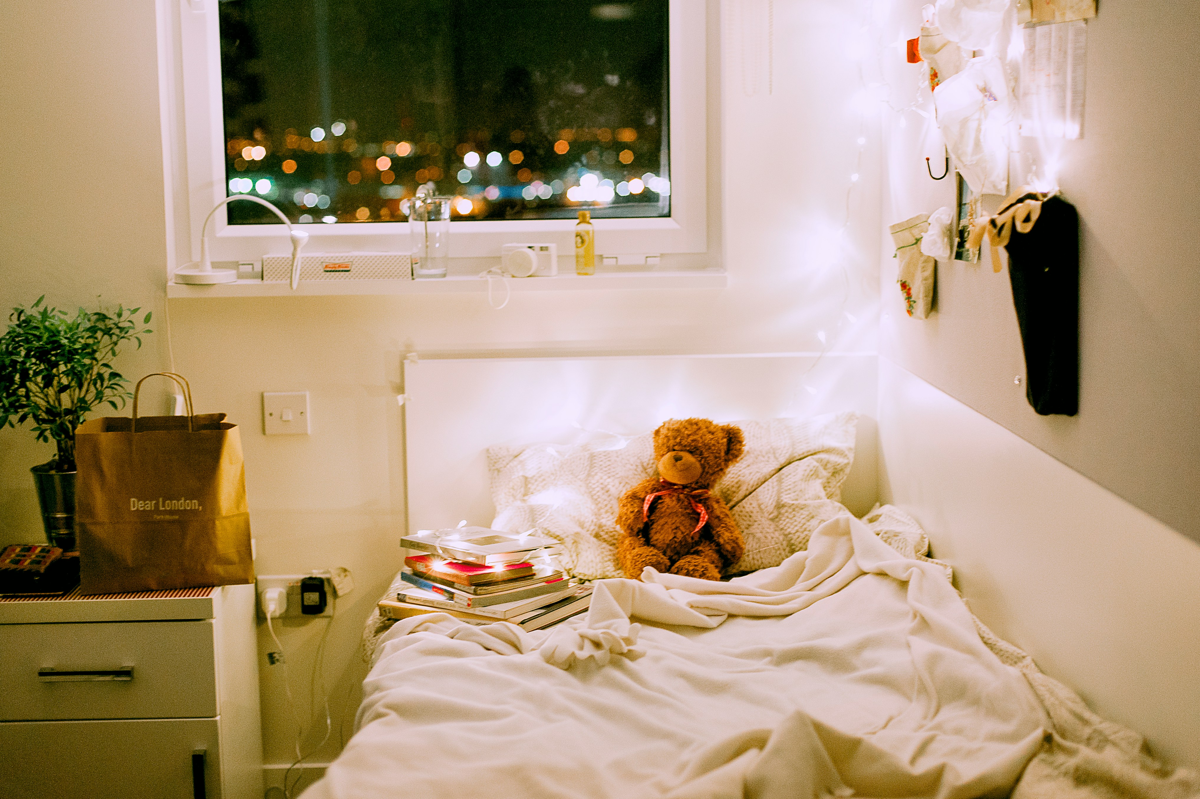 A single bed is well lit by several lamps. A bear and books are by the pillows. A window overlooking a city at night is behind the bed.